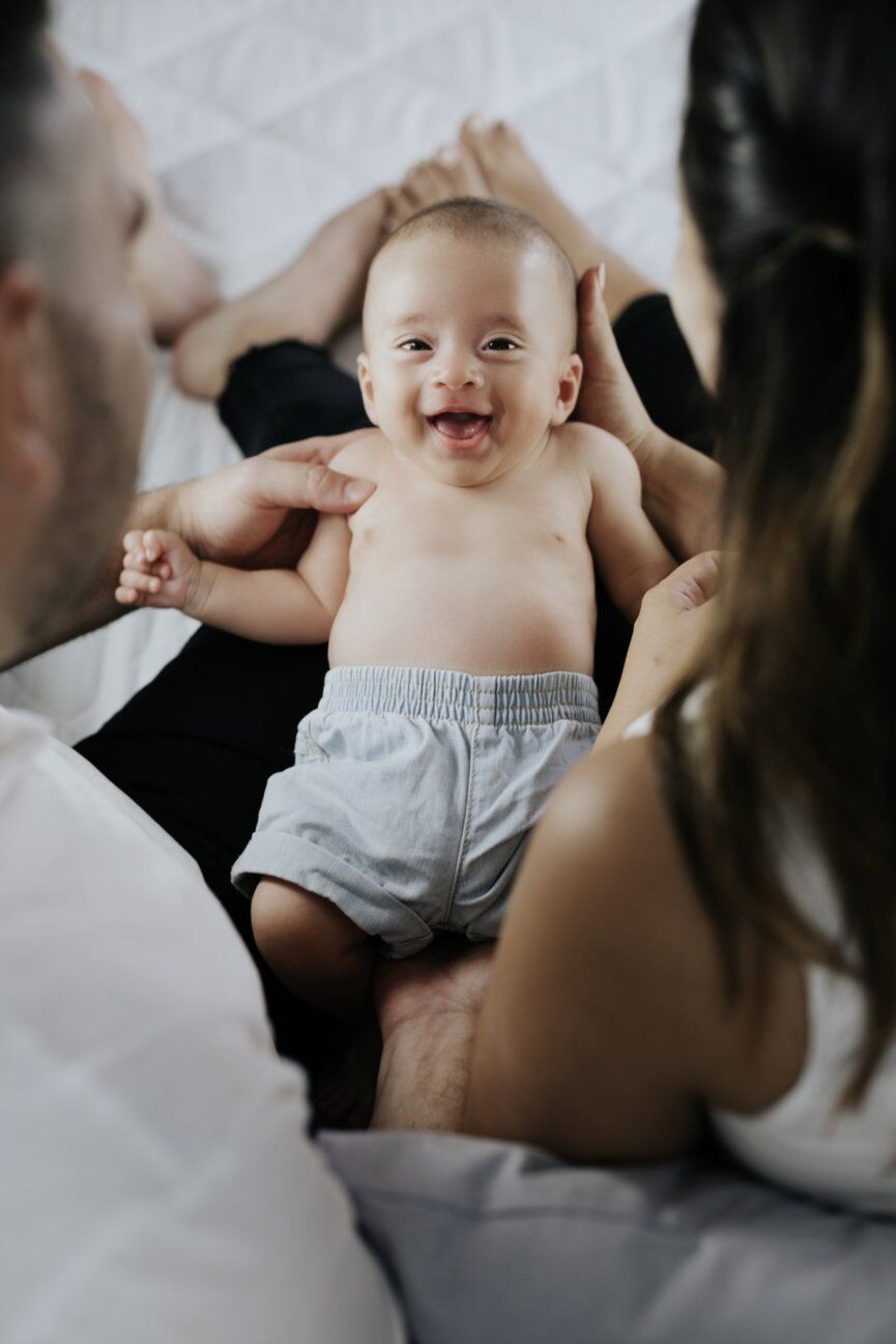 Neonato sorridente sdraiato sulle gambe di mamma e papà su un letto bianco, foto neonato a casa