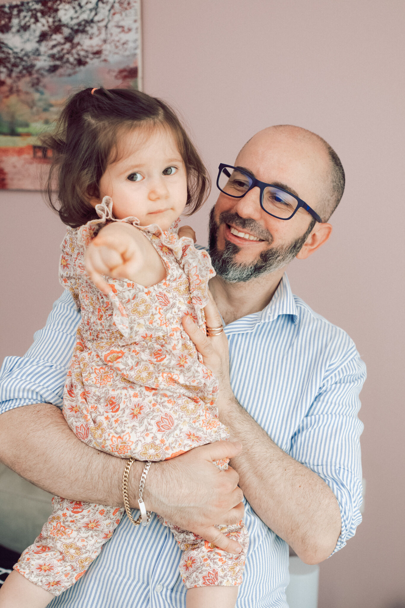 Papà con occhiali sorride tenendo in braccio la figlia con tutina a fiori che indica, foto di famiglia in casa