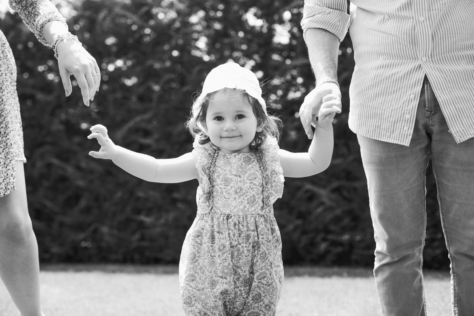 Bimba con cappellino muove i primi passi tenuta per mano dai genitori in giardino, foto in bianco e nero