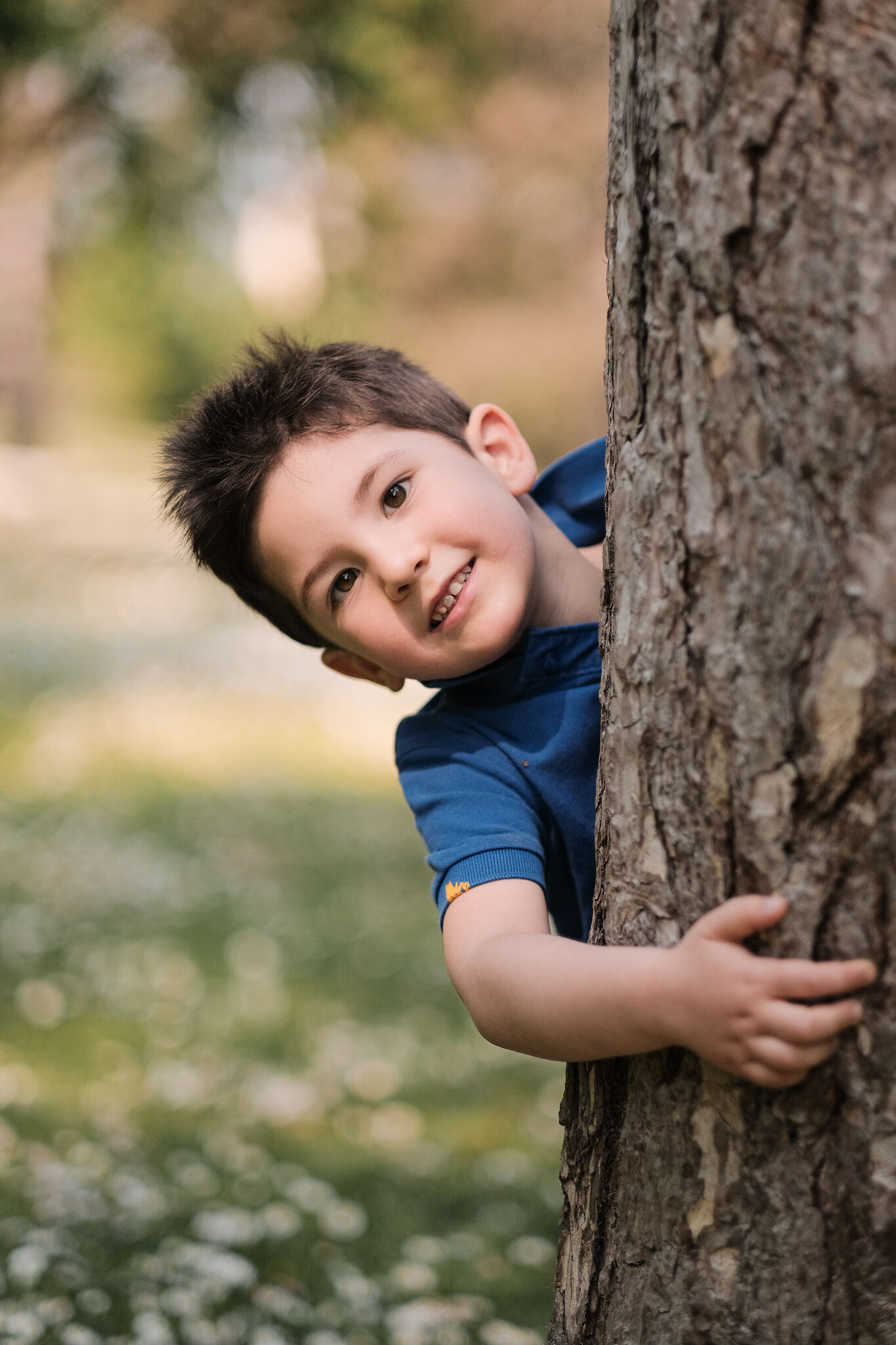 Bambino con maglietta blu che sbuca sorridendo da dietro un albero