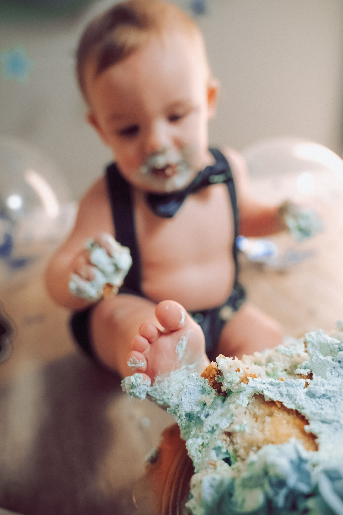 Piedino in primo piano affonda nella torta distrutta mentre il bimbo sorride sfocato sullo sfondo, smash cake