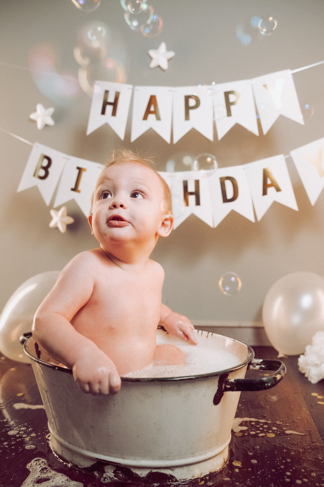Bimbo nella tinozza con la schiuma guarda le bolle di sapone sotto il festone di compleanno, luce calda