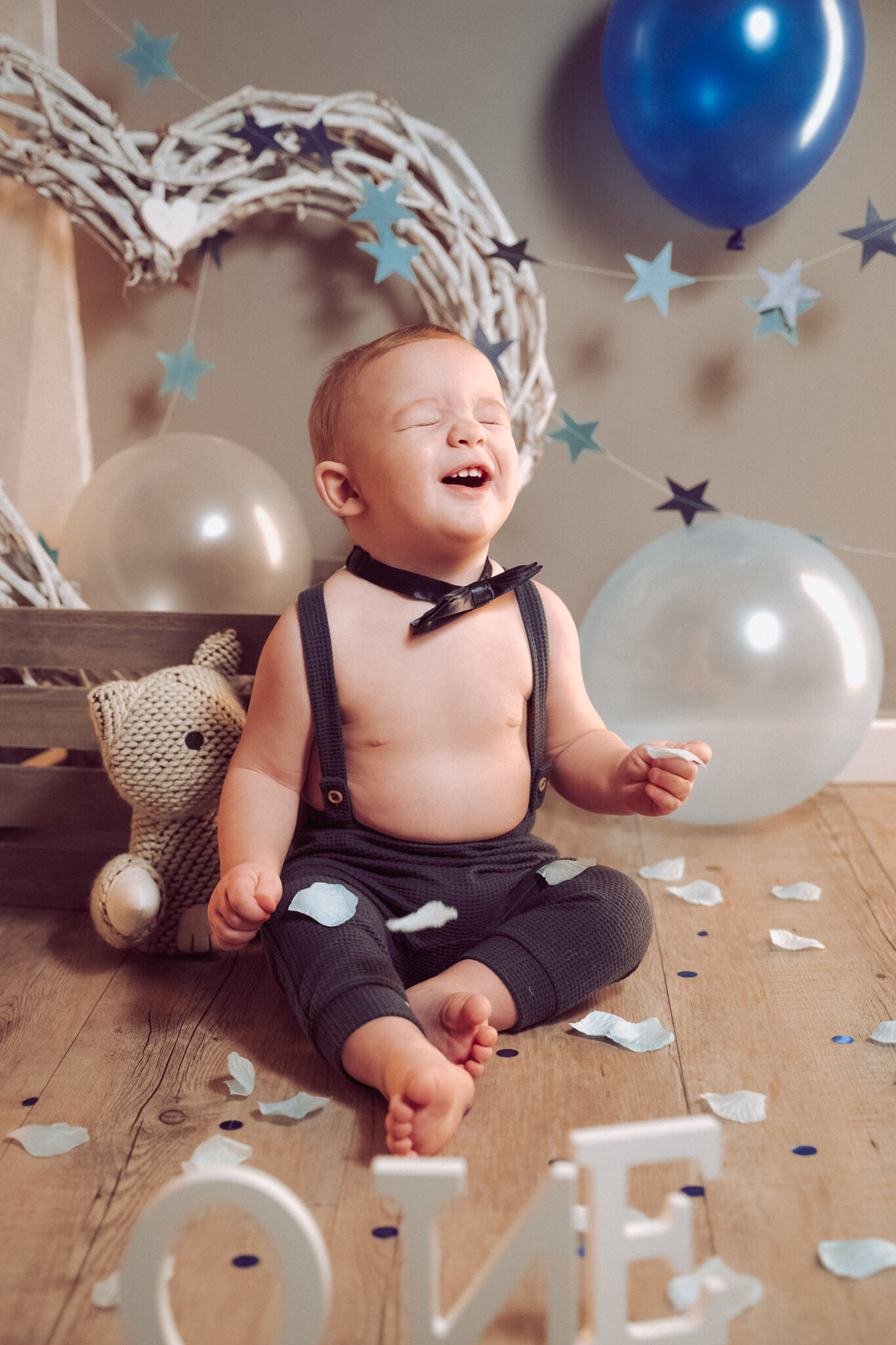 Bimbo con bretelle ride a occhi chiusi tra stelline blu, cuore di vimini e palloncini, primo compleanno in studio