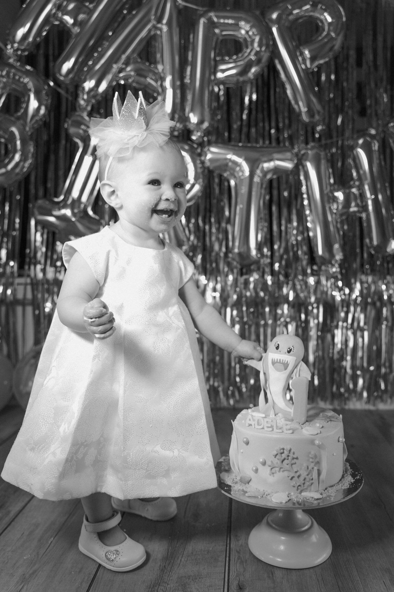 Bianco e nero di una bimba in abito bianco che ride accanto alla torta con squaletto, palloncini Happy Birthday