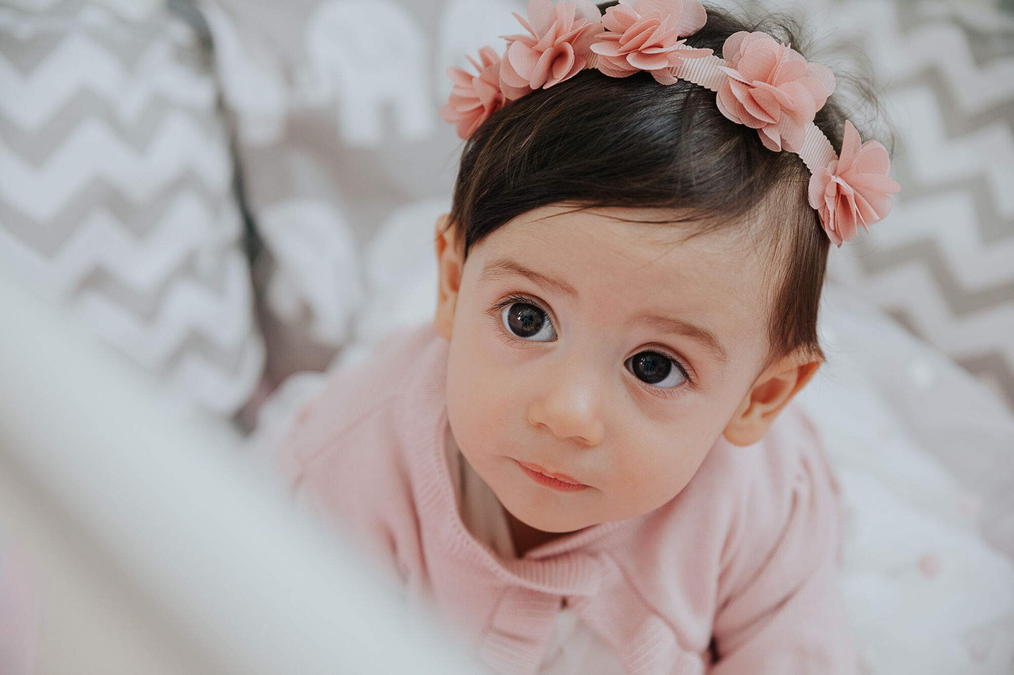 Bimba dagli occhi scuri con fascia di fiori rosa guarda in alto dal lettino, battesimo