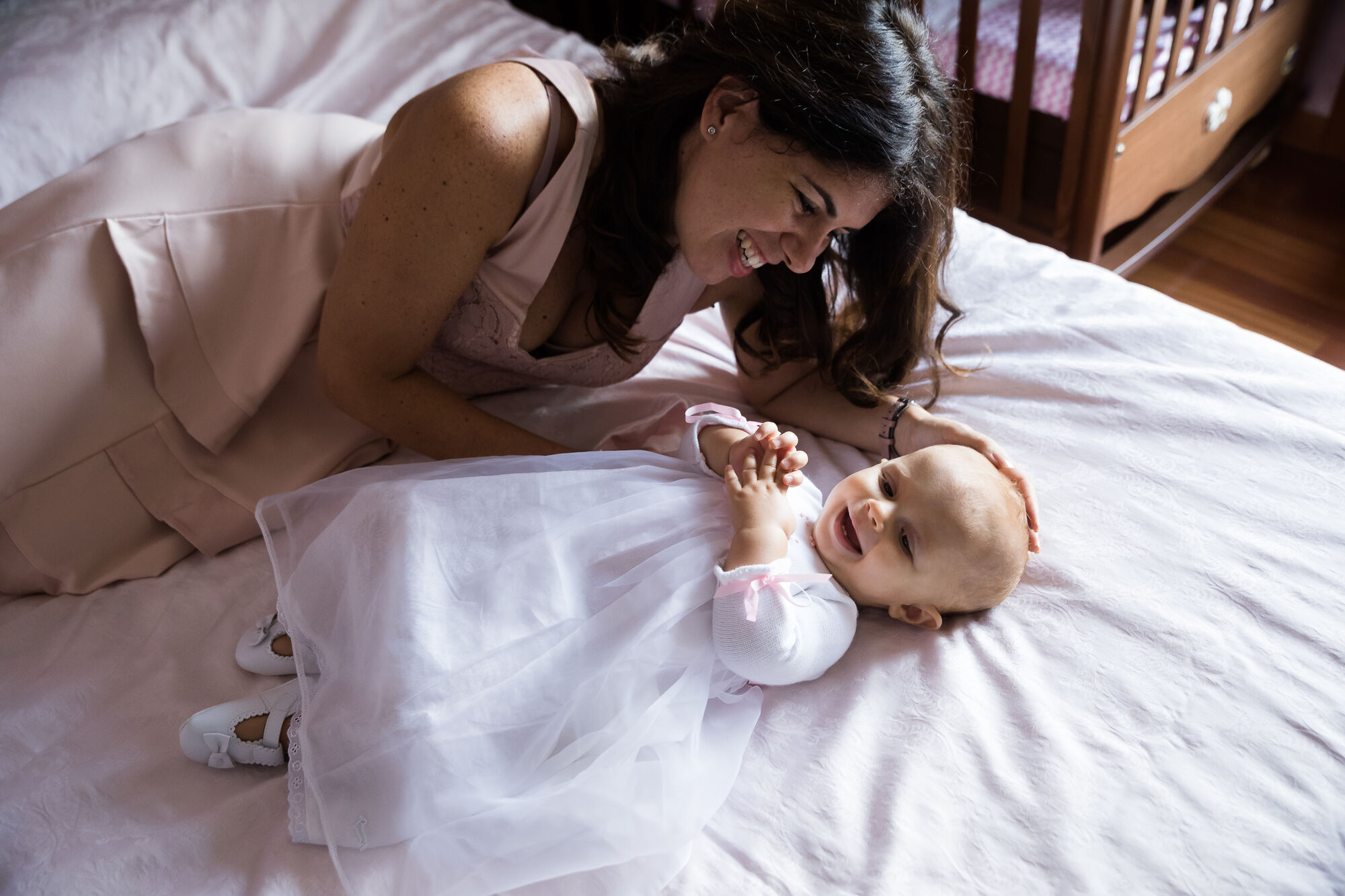 Mamma sorride chinata sul letto alla neonata in abito di tulle bianco, giorno del battesimo