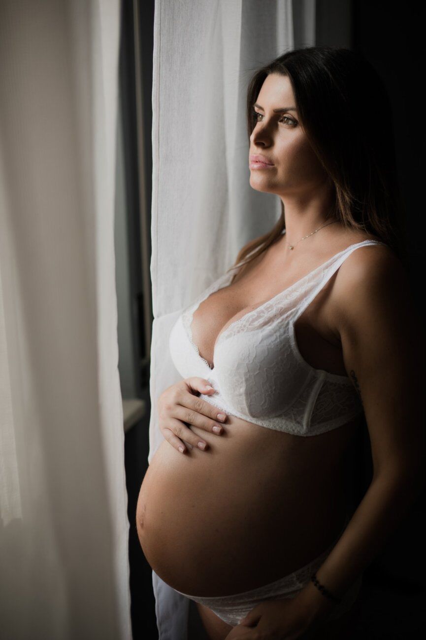Futura mamma in lingerie bianca accanto alla finestra, luce soffusa sul pancione e sguardo lontano