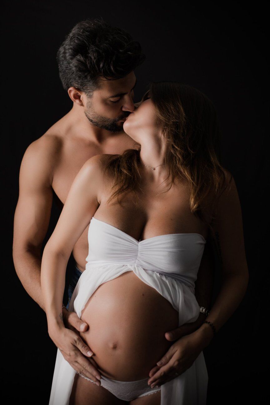 Bacio di coppia su fondo nero, lui abbraccia da dietro il pancione velato di bianco, servizio fotografico gravidanza