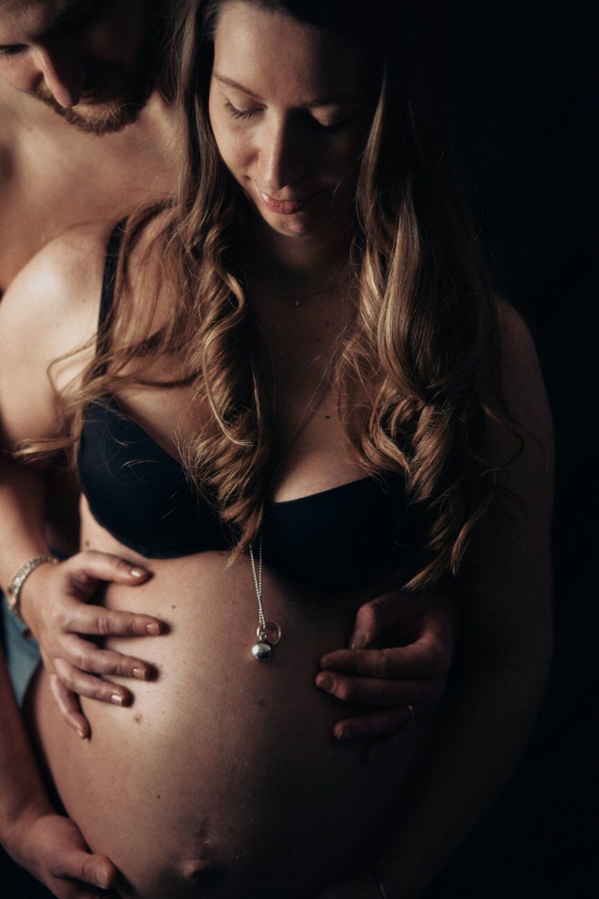 Lui abbraccia da dietro la compagna incinta in reggiseno nero, luce calda radente su capelli e pancione
