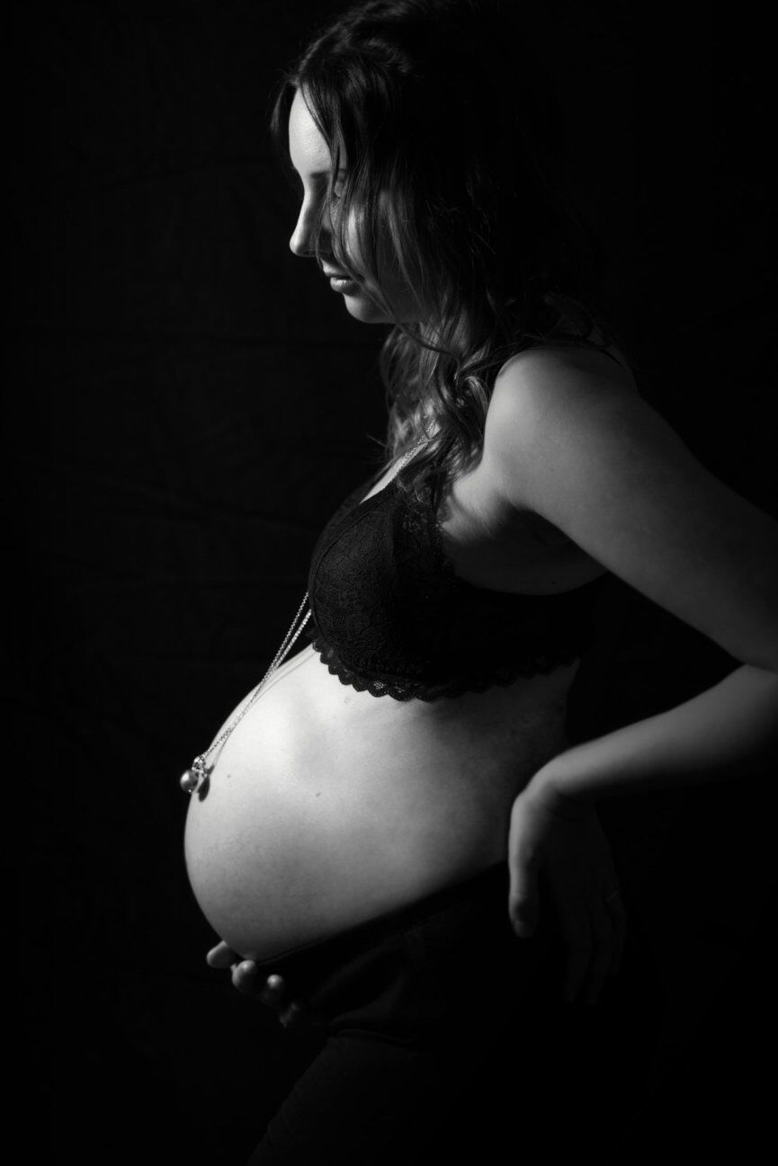 Profilo in chiaroscuro di donna incinta in reggiseno di pizzo nero con ciondolo sul pancione, bianco e nero