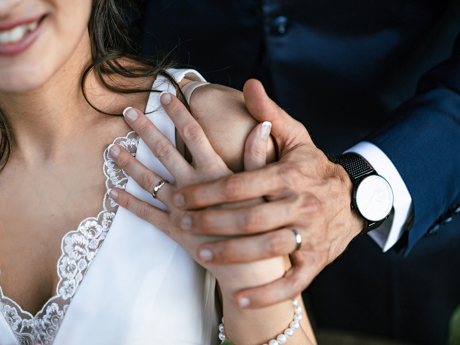 Mani dello sposo con orologio stringono quelle della sposa sorridente, dettaglio delle fedi nuziali