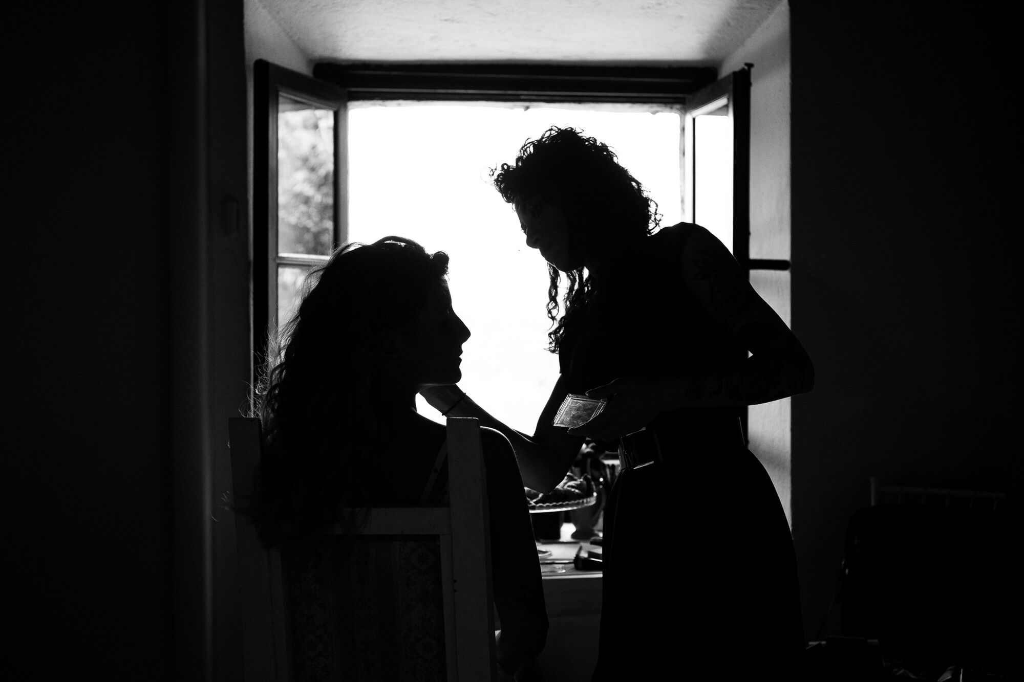 Truccatrice in controluce sistema il viso della sposa vicino alla finestra, getting ready del matrimonio