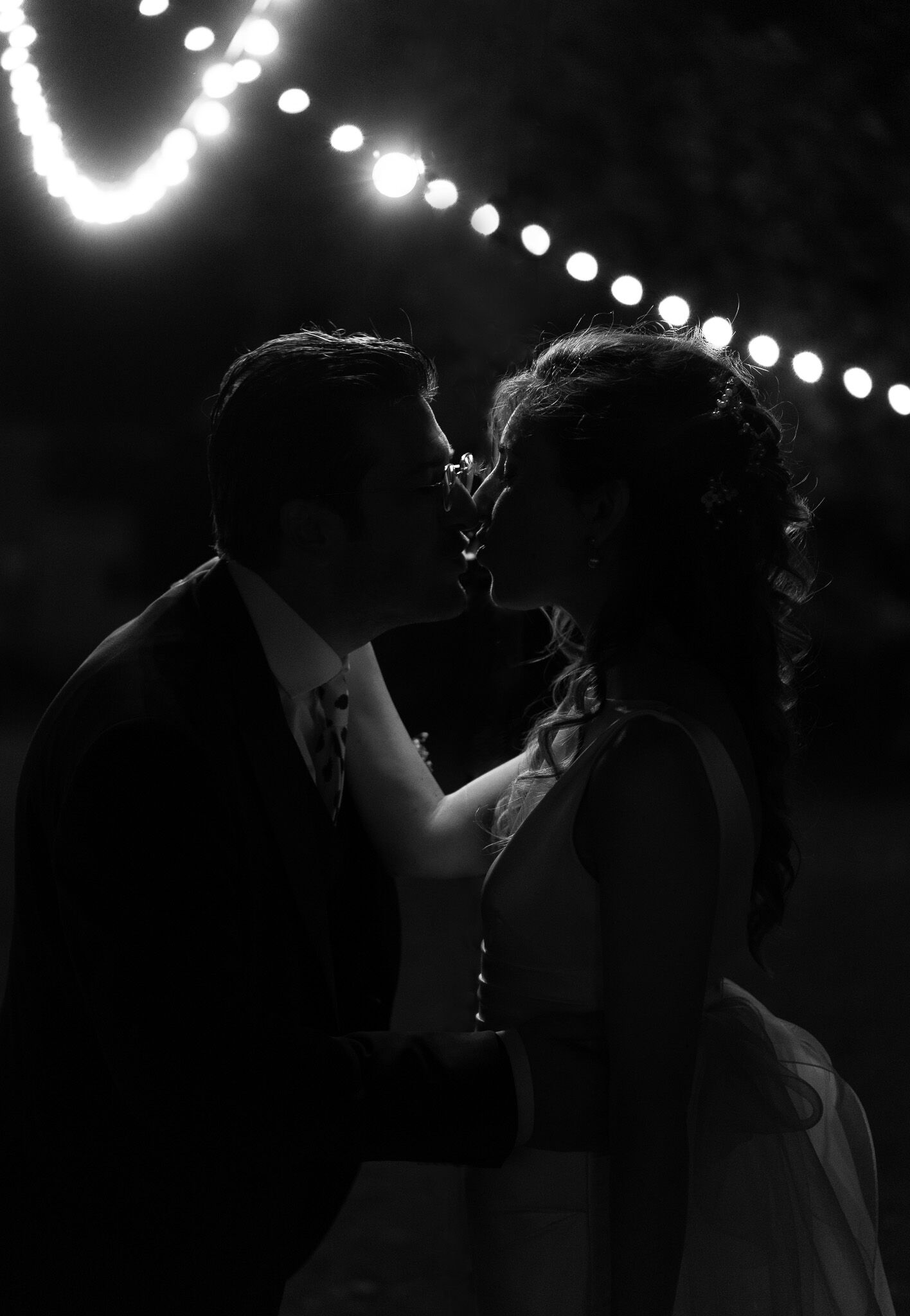 Bacio notturno degli sposi sotto una ghirlanda di lampadine, foto di matrimonio in bianco e nero
