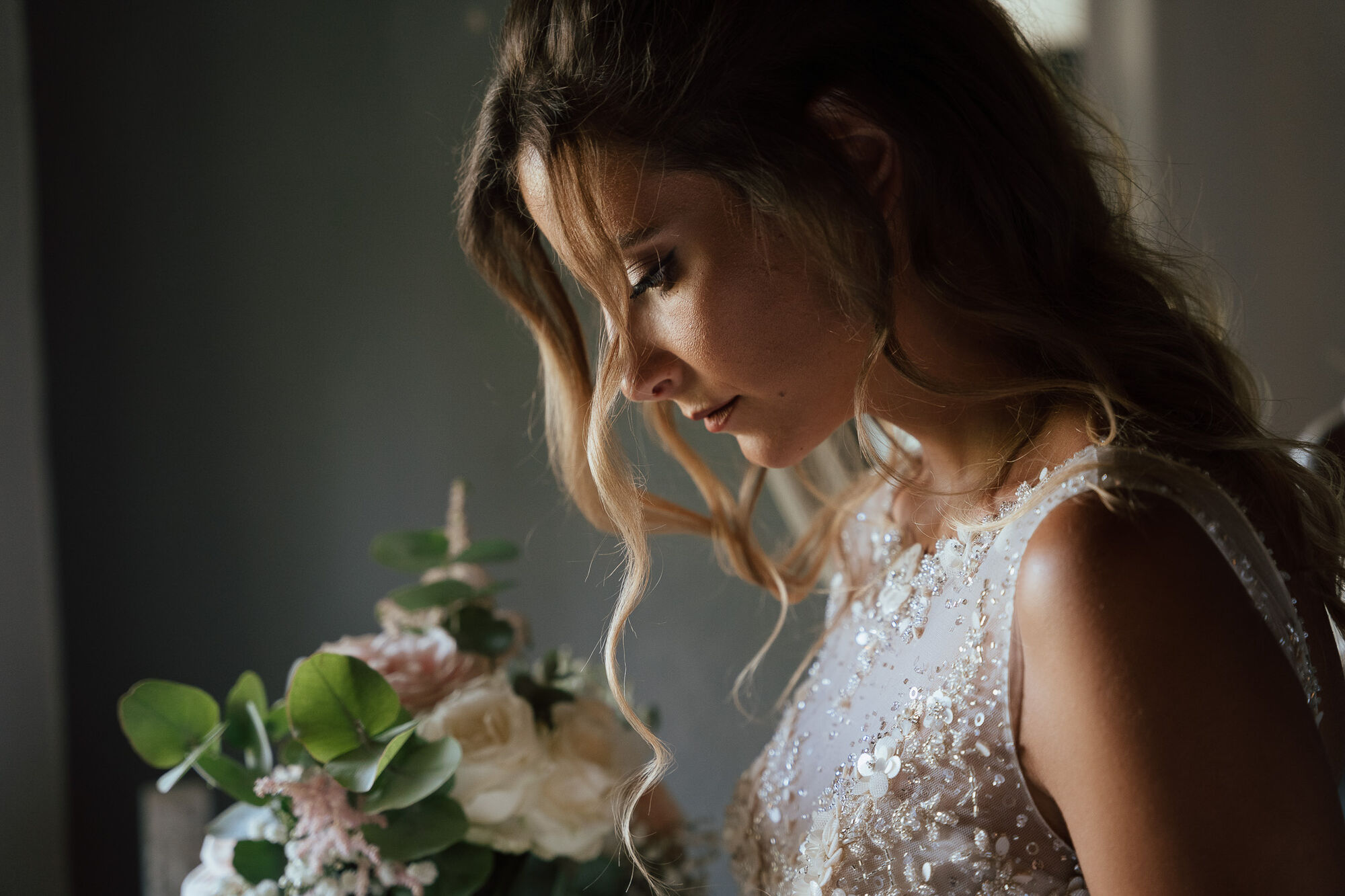 Sposa pensierosa con capelli mossi accanto al bouquet di rose rosa, luce morbida da finestra