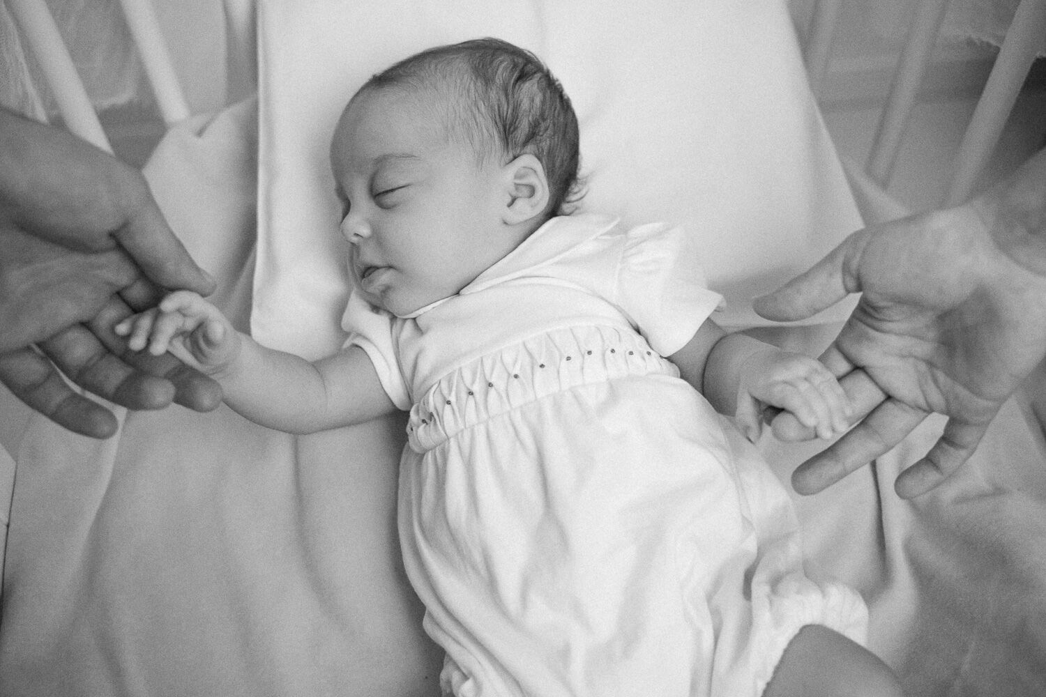 Neonato in tutina bianca dorme stringendo le dita dei genitori, foto neonato a casa in bianco e nero